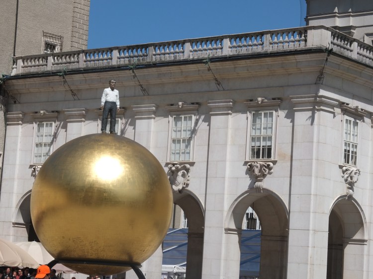 Das Bild zeigt das Kunstwerk von Stephan Balkenhohl "Sphära". Ein Mann steht auf der goldenen Kugel.