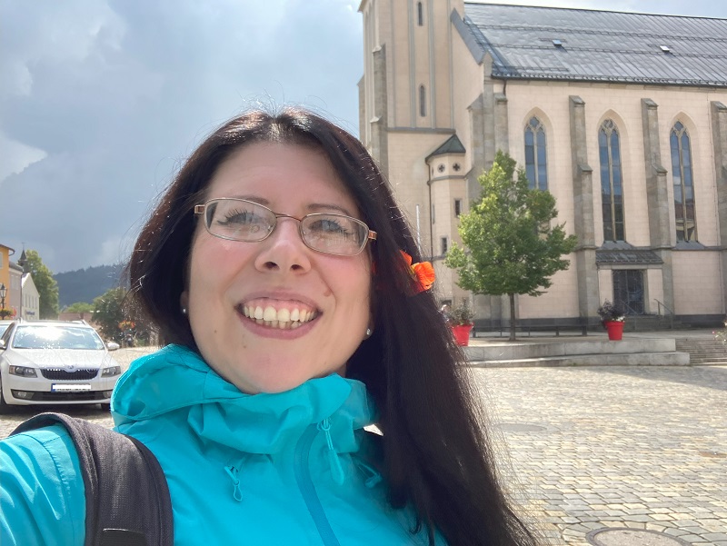 A young smiling woman with a flower in her long black hairs, wearing a light blue jacket.
