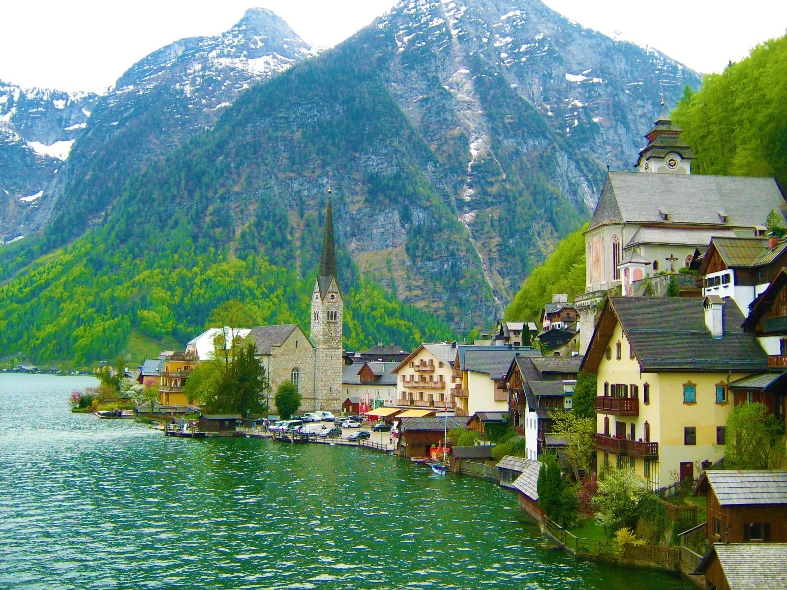 A little romantical austrian village with a name Hallstatt with a church with a long church tower on a green looking lake, on tha background are bluish high mountains.